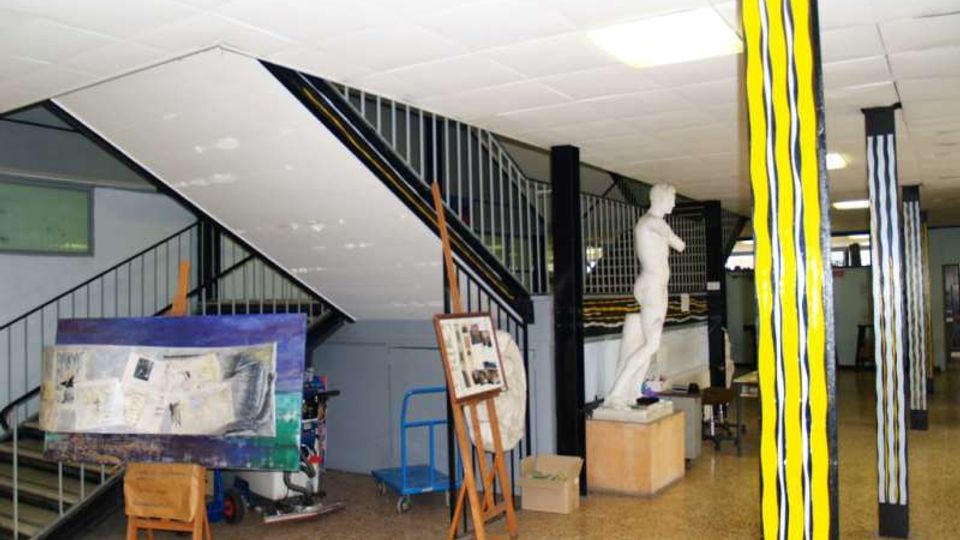 Atrio interno con scala metallica a vista, soffitto a pannelli e pavimento in graniglia, con colonne dipinte a strisce gialle e nere. Nello spazio sono presenti cavalletti con quadri e una statua su piedistallo, in un ambiente tipo scuola o centro culturale.
