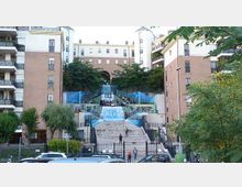 Grande scalinata urbana tra edifici residenziali in mattoni e intonaco, con terrazze laterali e rampe simmetriche. Al centro si vedono gradoni e strutture con teli blu simili a cascate, circondati da alberi e cancellate in ferro, con strada e auto in primo piano.