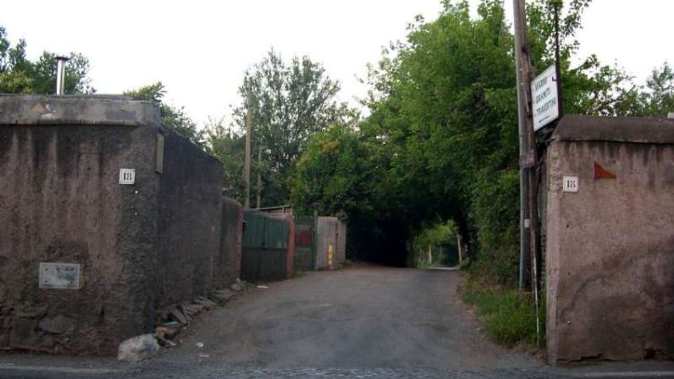 Ingresso a una strada stretta asfaltata, con pavimentazione in sampietrini in primo piano e due muri in cemento ai lati con numeri civici e segnaletica. Sullo sfondo la via prosegue tra vegetazione fitta e alberi, con cancelli e recinzioni lungo i lati.