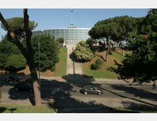 Edificio moderno a pianta semicircolare con ampia facciata vetrata, situato in cima a una collina. Una lunga scalinata centrale attraversa un prato alberato e conduce all’ingresso, con una strada trafficata in primo piano.