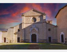 Facciata di una chiesa in pietra su una piccola piazza, con grande finestra circolare centrale e portale monumentale incorniciato. Ai lati si vedono edifici adiacenti e un cielo al tramonto con nuvole rossastre.