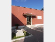 Ingresso di un edificio basso con facciata intonacata color rosso mattone, piccola pensilina e gradini in marmo che conducono a una porta vetrata con inferriata. Davanti ci sono un vialetto pavimentato, una piccola aiuola con blocco in pietra e una targa informativa sulla parete.