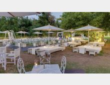 Area lounge all’aperto in un giardino con divanetti e tavolini bianchi, ombrelloni grandi e sedute in ferro battuto. Sullo sfondo si vedono una pedana con molte sedie disposte in file e alberi fitti che circondano lo spazio.