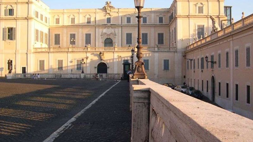 Ampio cortile pavimentato in pietra davanti a un grande palazzo storico color crema con facciata simmetrica, finestre con persiane e un portale centrale ad arco. In primo piano si vede un parapetto in pietra con lampione in stile antico lungo una rampa, con un edificio laterale sul lato destro.