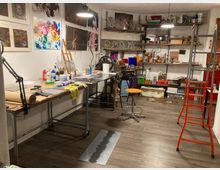 Interno di uno studio d’arte con grandi tavoli da lavoro su ruote, illuminazione a sospensione e pareti coperte da stampe e dipinti. Scaffalature sul fondo con materiali e strumenti artistici, sgabello centrale e scala rossa a destra su pavimento in legno scuro.