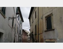 Stretta via di un centro storico tra edifici alti in muratura con facciate intonacate e persiane scure, illuminate da lanterne a muro. Cavi sospesi attraversano la strada sotto un cielo nuvoloso.