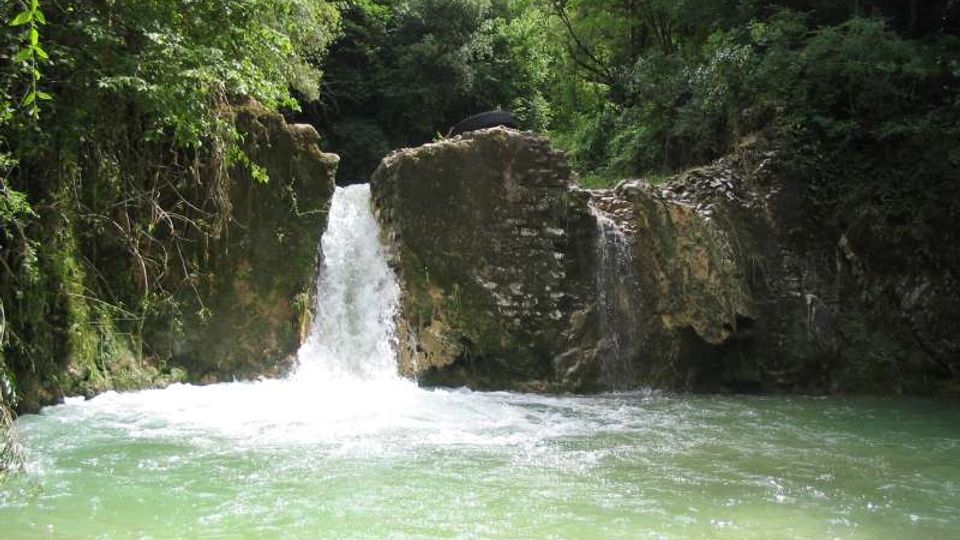 Piccola cascata che scende tra rocce e muri in pietra in una pozza d’acqua verde-azzurra. Intorno si vede una fitta vegetazione di alberi e arbusti che circonda il corso d’acqua.