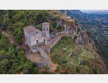 Chiesetta in pietra con campanile e tetto in tegole, costruita su un crinale roccioso con terrazze e una scalinata in muratura. Intorno ci sono boschi e pareti scoscese, con vista sulla valle e sui campi sottostanti.