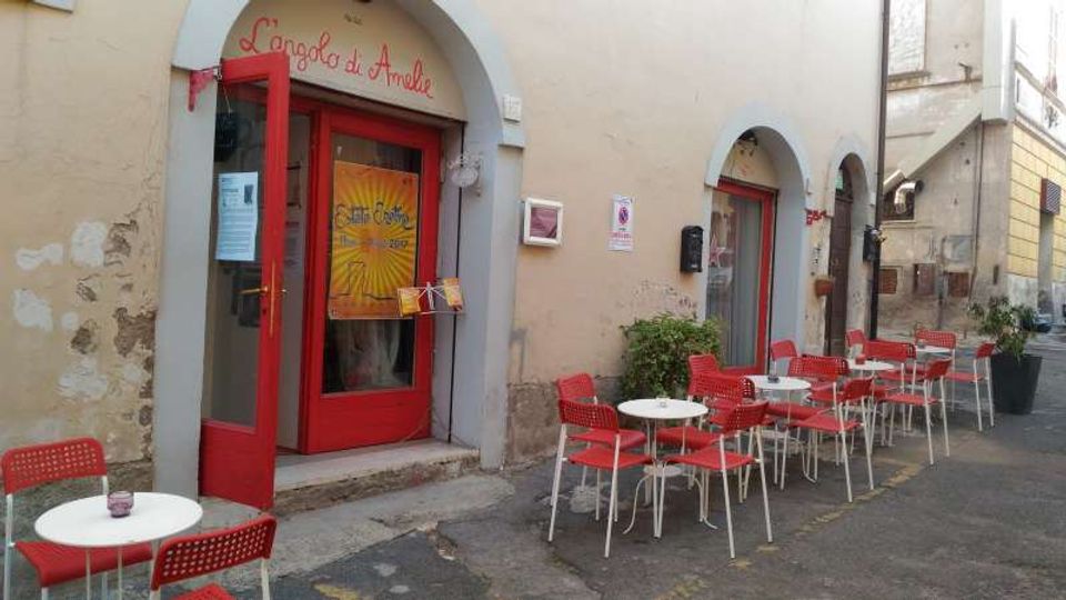 Facciata di un piccolo bar in una strada stretta, con portone e infissi rossi sotto un arco e pareti intonacate. Davanti all’ingresso ci sono tavolini rotondi bianchi con sedie rosse disposti lungo il marciapiede, accanto a finestre ad arco.