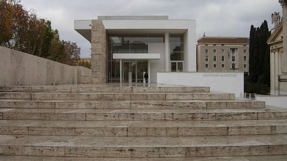 Edificio moderno con facciata vetrata e volume bianco sporgente, raggiungibile tramite un’ampia scalinata in pietra. Ai lati si vedono muri in travertino, alberi e altri edifici in un contesto urbano sotto cielo nuvoloso.