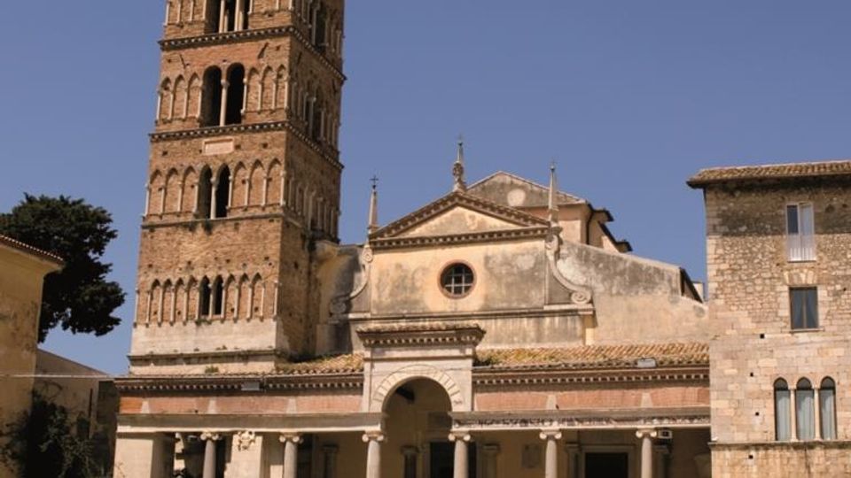 Chiesa storica in pietra con ampia scalinata e portico colonnato, affiancata da un alto campanile in mattoni con arcate su più livelli. Davanti si apre una piazza lastricata, con edifici in pietra ai lati sotto un cielo sereno.