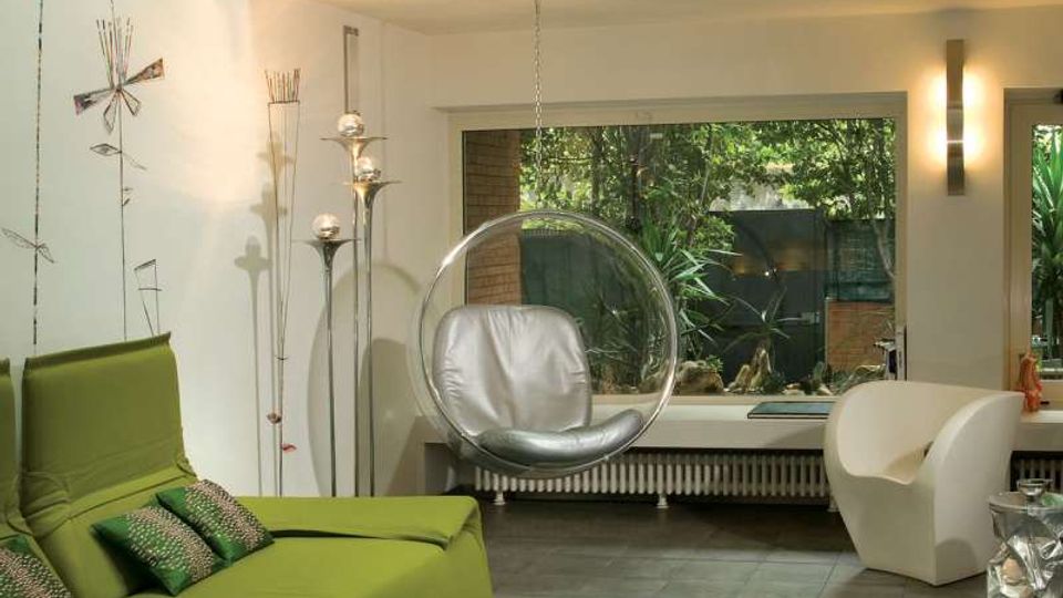 Soggiorno moderno a doppia altezza con soppalco e ringhiera in metallo, pareti bianche e pavimento in piastrelle scure. Divano verde, tavolino bianco su tappeto, poltrone di design e ampie finestre che affacciano su un giardino esterno.