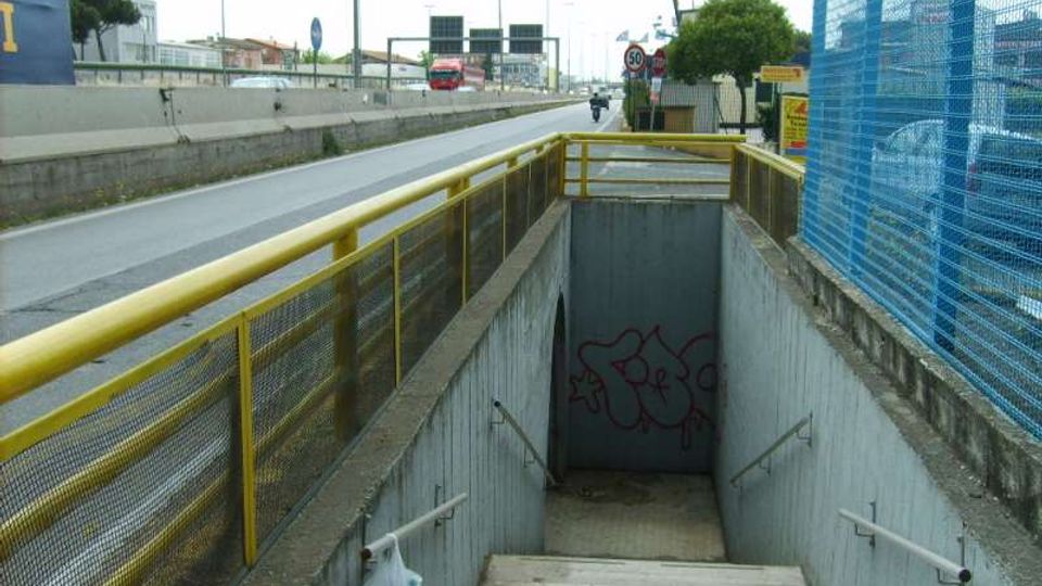 Sottopassaggio pedonale con scala in cemento che scende sotto una strada a scorrimento veloce, affiancata da una ringhiera gialla e una recinzione metallica blu. In fondo alla rampa si vede un muro con graffiti, mentre lungo la carreggiata ci sono cartelli stradali e infrastrutture urbane.