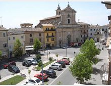 Piazza cittadina con parcheggi, aiuole e alberi, circondata da edifici a più piani con tetti in coppi. Sullo sfondo domina una grande chiesa in pietra con facciata classica, campanile e portale centrale.