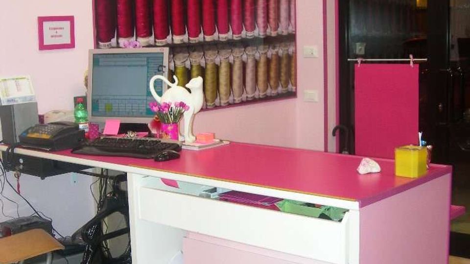 Interno di un negozio con pareti rosa e banco reception bianco con piano fucsia, su cui sono presenti computer e piccoli oggetti. Sulla parete dietro il banco c’è un grande pannello con rocchetti di filo colorati disposti in file, e a destra si vede una porta vetrata scorrevole.
