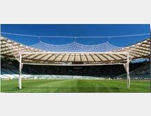 Vista panoramica di uno stadio all’aperto con ampio campo da calcio in erba e gradinate circolari. La copertura curva è sostenuta da pilastri e da una struttura a rete di cavi, con cielo sereno sullo sfondo.
