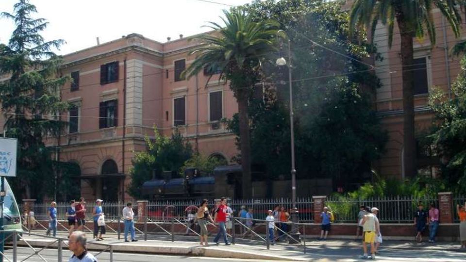 Edificio storico a più piani con facciata rosa e finestre scure, affacciato su una strada con recinzione metallica e marciapiede. Davanti all’edificio ci sono palme e alberi alti, con lampione e cavi sospesi nell’area urbana circostante.