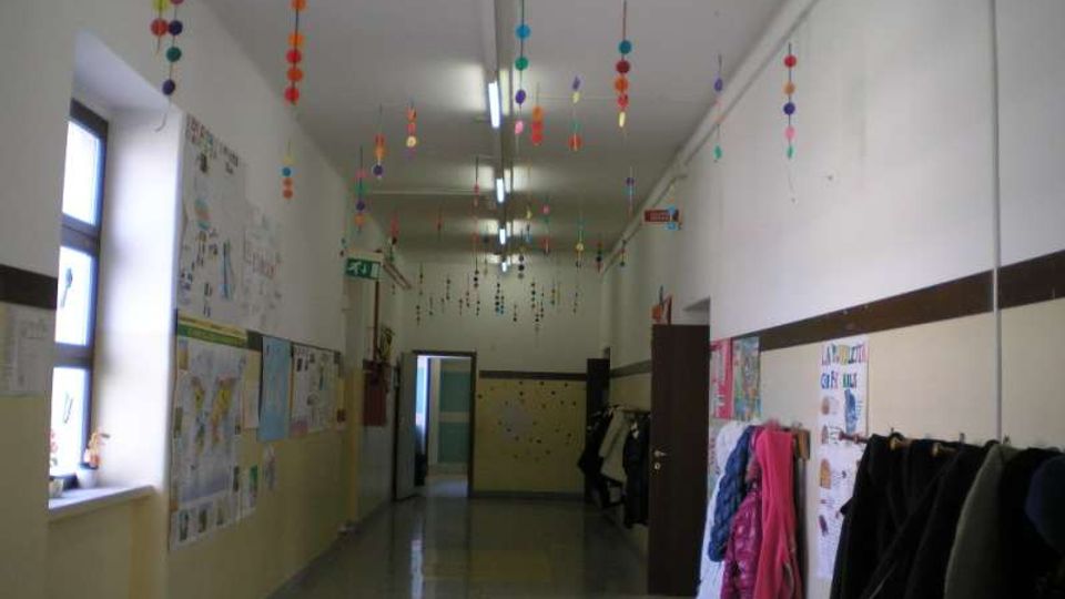 Corridoio interno di una scuola con pavimento piastrellato lucido, pareti bianche con fascia beige e disegni e cartelloni appesi. Dal soffitto pendono decorazioni colorate e lungo le pareti ci sono finestre, armadietti e appendiabiti con giacche; in fondo si vede una porta aperta.