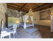 Camera da letto in stile rustico con soffitto a travi in legno, pareti in pietra a vista e pavimento in cotto. Al centro c’è un letto a baldacchino bianco con tende, illuminato da abat-jour e da un lampadario, con una piccola scrivania e sedia bianche sul lato.
