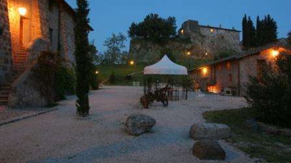 Cortile di un casale in pietra al crepuscolo con vialetti di ghiaia, luci esterne calde e un gazebo bianco al centro. Sullo sfondo si vedono colline alberate, cipressi e un edificio in pietra su un’altura.