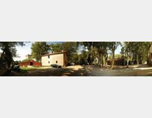 Cortile sterrato ombreggiato da alti alberi, con una piccola casa rurale intonacata al centro e altri edifici bassi sullo sfondo. Sul lato sinistro si vede un muro rosso con un arco d’ingresso e, nell’area, alcuni veicoli parcheggiati lungo i bordi del piazzale.