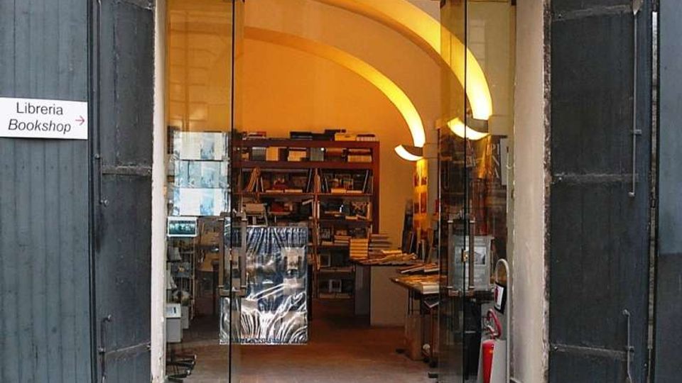 Ingresso di una libreria con grandi porte scure aperte e vetrate scorrevoli, che conduce a un interno con soffitto a volte illuminato e scaffali pieni di libri. A sinistra è visibile un cartello “Libreria/Bookshop” che indica l’entrata.