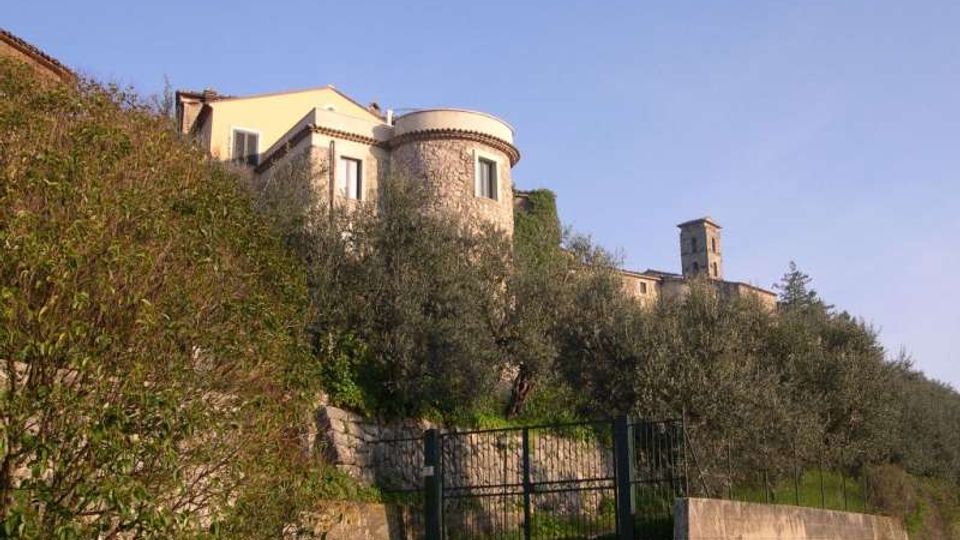 Edificio in pietra su un pendio collinare, con torretta cilindrica e finestre, circondato da ulivi e vegetazione. In primo piano si vedono un cancello metallico e muri di contenimento in pietra lungo una strada.