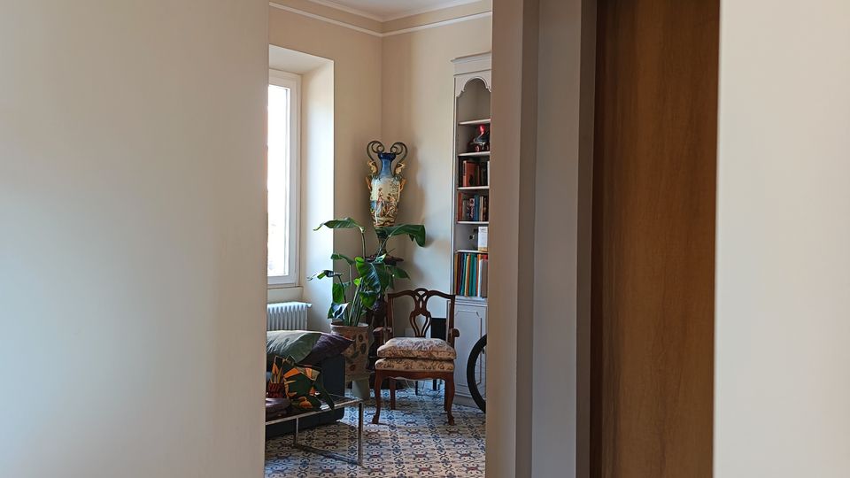 Vista da un corridoio verso un salotto con pavimento in piastrelle decorative a motivi geometrici. Si vedono una grande finestra, una libreria bianca, sedie imbottite, piante in vaso e una lanterna a sospensione, con una porta in legno sulla destra.