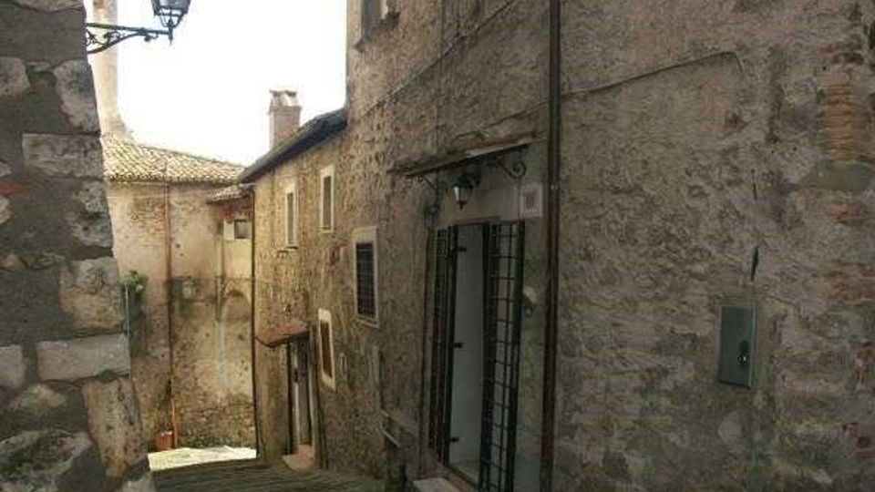 Vicolo stretto in discesa con gradini in pietra tra edifici in muratura di pietra e mattoni, con porte e finestre dai telai scuri. Un lampione a parete e cavi esterni corrono lungo le facciate, in un contesto di borgo storico.