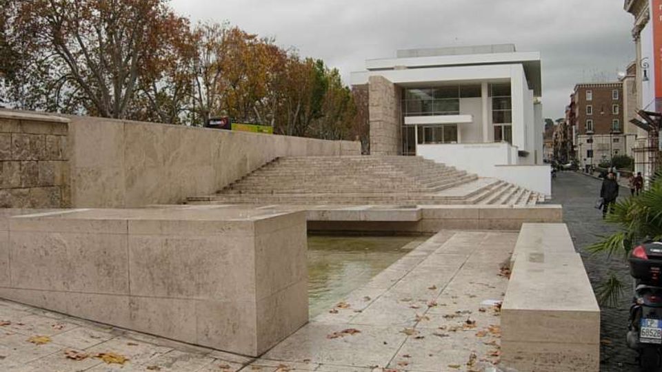 Spazio urbano con ampia scalinata e piattaforme in pietra chiara che circondano una vasca d’acqua bassa. Sullo sfondo si vede un edificio moderno bianco con grandi vetrate, affiancato da alberi e da una strada cittadina.
