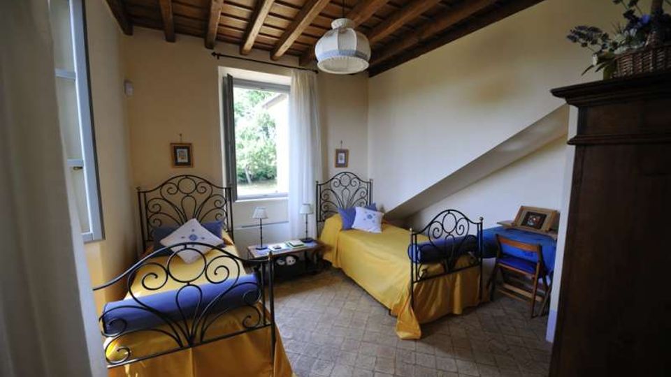 Camera da letto con due letti singoli in ferro battuto, copriletti gialli e cuscini blu, con finestra centrale e piccolo tavolino tra i letti. Soffitto in legno a travi a vista, pavimento in piastrelle e arredi essenziali tra cui una scrivania e un armadio scuro.