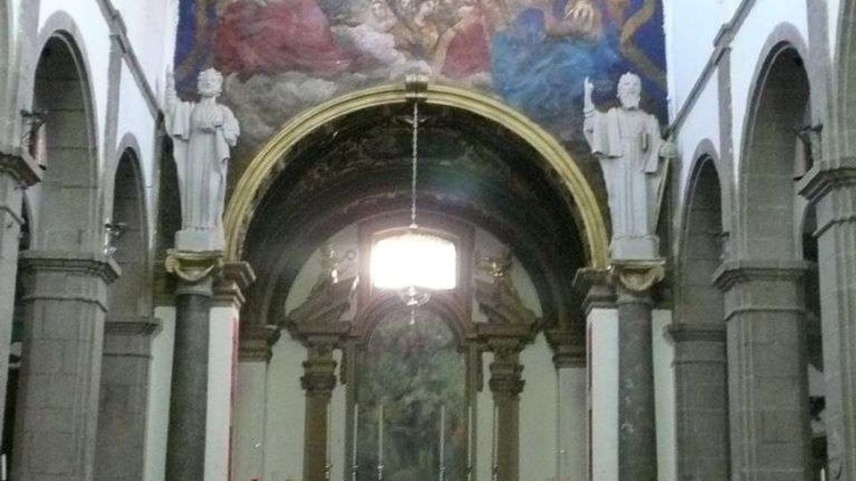 Interno di una chiesa con navata centrale fiancheggiata da arcate e colonne in pietra, che conduce all’altare maggiore rialzato. Sopra l’altare si vede un grande affresco semicircolare e statue su piedistalli ai lati dell’arco, con banchi in legno in primo piano.