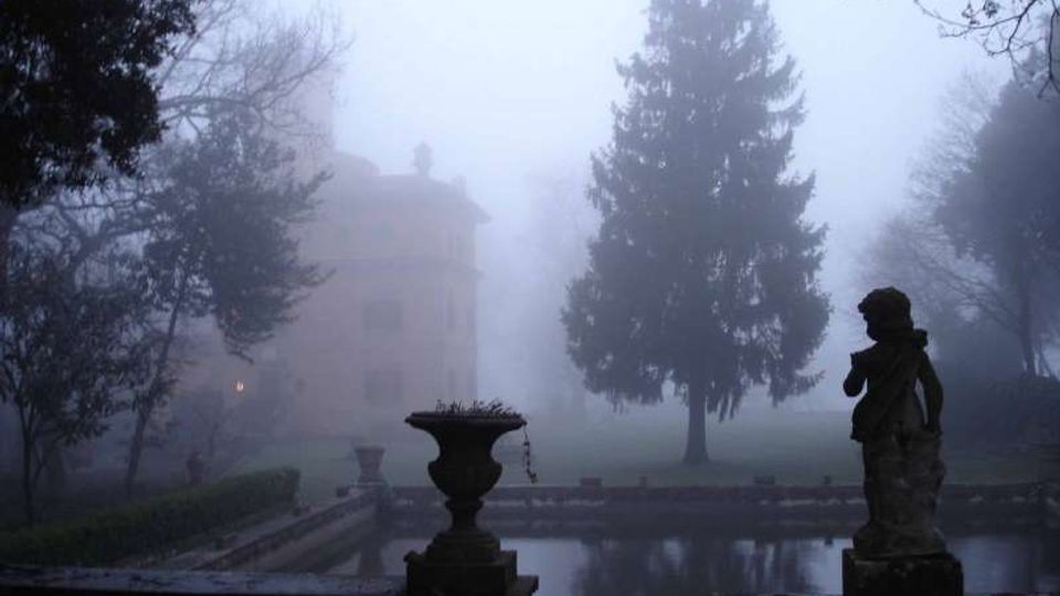 Giardino di una villa immerso nella nebbia, con una vasca rettangolare d’acqua in primo piano, un vaso ornamentale e una statua su piedistallo. Sullo sfondo si intravedono la facciata della villa e un grande albero sempreverde tra alberi spogli.