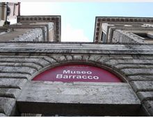 Ingresso del Museo Barracco visto dal basso, con arco in pietra bugnata e insegna rossa “Museo Barracco” sopra il varco. Ai lati si elevano due facciate in muratura con cornici e finestre, incorniciate dal cielo sullo sfondo.