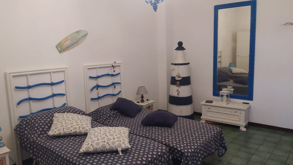 Camera da letto in stile marinaro con due letti singoli affiancati, testiere bianche con dettagli blu e biancheria scura a motivi. Sulla parete ci sono uno specchio con cornice blu e un mobile basso bianco, mentre nell’angolo si vede una decorazione a forma di faro; pavimento in piastrelle verdi e lampadario a soffitto.