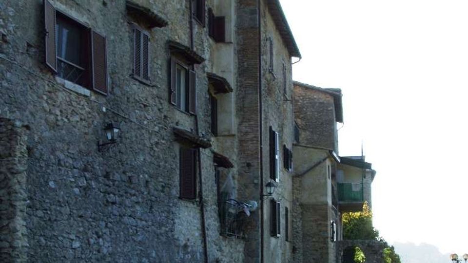 Fila di edifici storici in pietra con persiane scure e piccole finestre, affacciati su un prato lungo una stretta riva murata. Sul lato destro si intravede un parapetto in pietra e un tratto di panorama in lontananza.