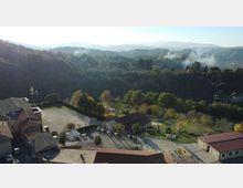 Veduta panoramica dall’alto di un piccolo centro con edifici dai tetti in laterizio, un piazzale e aree verdi alberate. Sullo sfondo si estendono colline e montagne boscose con leggere velature di nebbia o fumo tra le valli.