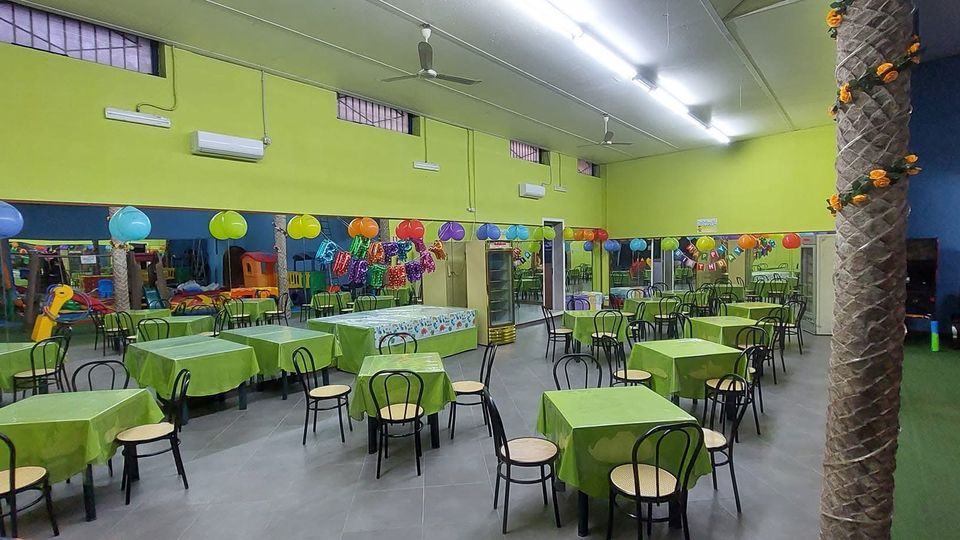Ampia sala interna per feste per bambini con pareti verde e blu, tavoli coperti da tovaglie verdi e sedie nere, illuminazione a neon e ventilatori a soffitto. Palloncini e decorazioni colorate sono appesi lungo la parete con specchi, con area giochi e strutture per bambini sullo sfondo.