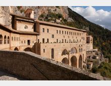 Grande complesso monastico in pietra chiara con arcate e loggiati, costruito a ridosso di una parete rocciosa e disposto su più livelli. Sullo sfondo si vedono il pendio boscoso della valle e il cielo parzialmente nuvoloso.