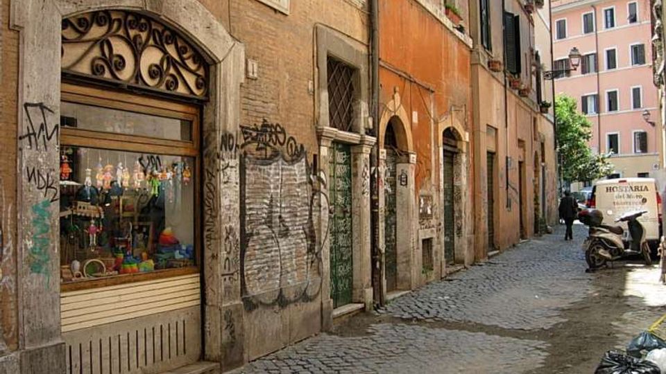 Vicolo stretto con pavimentazione in sampietrini, fiancheggiato da edifici storici intonacati con portoni ad arco, finestre con persiane e facciate segnate da graffiti. In primo piano si vede una vetrina con inferriata decorativa e, più avanti, un piccolo veicolo e sacchi della spazzatura lungo la strada.