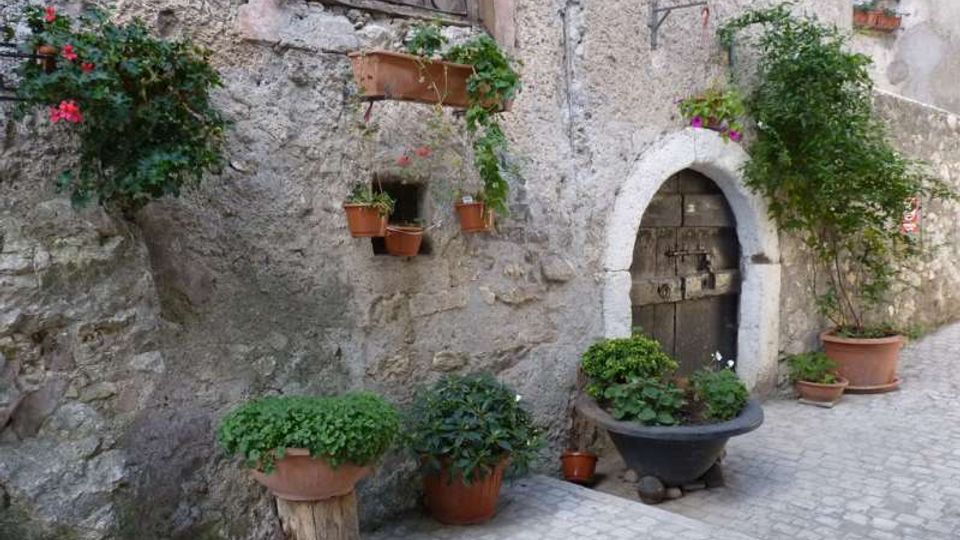 Facciata di una casa in pietra su una viuzza acciottolata, con porta ad arco in muratura e finestra con inferriate. Vasi e fioriere in terracotta con piante e fiori decorano il muro e il piccolo spazio davanti all’ingresso.