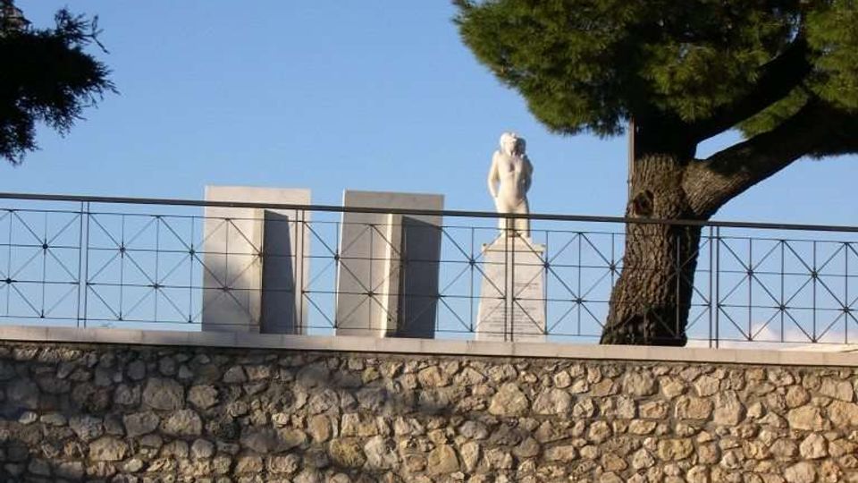 Terrazza rialzata con muri in pietra e ringhiera metallica, con una statua su piedistallo e grandi blocchi rettangolari sullo sfondo. Un grande albero sempreverde domina il lato destro contro un cielo sereno.