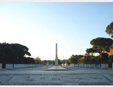 Ampia piazza monumentale pavimentata con decorazioni geometriche e figurative, delimitata da bassi parapetti e alberi ai lati. Al centro si erge un alto obelisco in pietra lungo un asse prospettico verso l’orizzonte.
