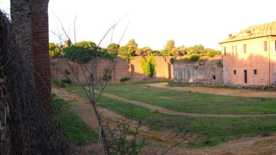 Cortile erboso con sentieri di terra curvi, circondato da alte mura in mattoni e vegetazione sullo sfondo. A destra si vede un edificio color ocra con piccole finestre, mentre in primo piano compaiono tronchi e rami secchi.