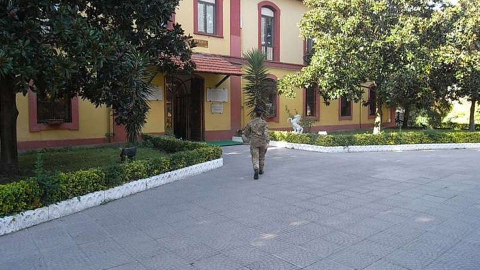 Edificio a due piani in stile tradizionale, con facciata gialla e bordature rosse attorno a finestre e ingressi, affacciato su un ampio piazzale pavimentato. Davanti si trovano aiuole bordate di bianco con siepi e alberi, che incorniciano il percorso verso l’entrata.
