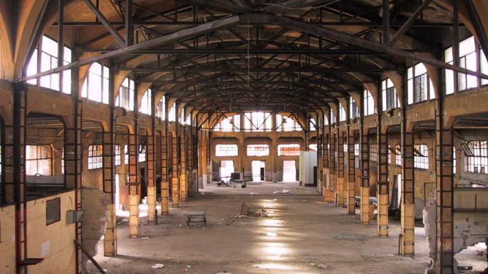 Interno di un grande capannone industriale abbandonato, con ampia navata centrale e struttura a capriate metalliche a vista sotto il tetto. Lungo le pareti si vedono finestre alte e pilastri in muratura, con pavimento in cemento parzialmente coperto da detriti e aperture sul fondo.