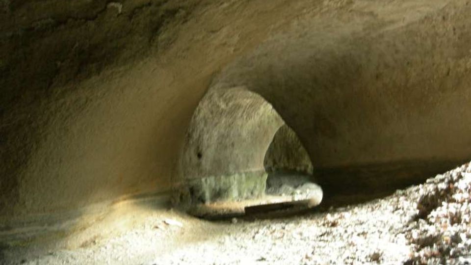 Interno di una grotta o tunnel scavato nella roccia, con volta arrotondata e pareti grezze. In fondo si vede un piccolo passaggio ad arco illuminato, con pavimento terroso e detriti.