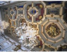 Soffitto interno riccamente decorato con cassettoni ottagonali e riquadri geometrici in rilievo, con dettagli dorati e inserti colorati. Lampadari in vetro sospesi illuminano l’ambiente, evidenziando le cornici e gli elementi ornamentali del soffitto.