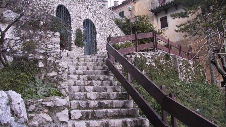 Scalinata in pietra che sale tra muri di roccia verso un edificio in pietra con due finestre ad arco protette da grate. A destra corre una passerella con parapetto in legno, circondata da vegetazione e case con balconi sullo sfondo.