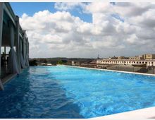 Piscina a sfioro su una terrazza panoramica, con acqua azzurra e bordo bianco affacciato su un paesaggio urbano di edifici bassi. A sinistra si vede una struttura coperta con tende leggere, sotto un cielo ampio con nuvole.